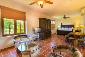 Une chambre à coucher comprenant un lit, un téléviseur, une armoire en bois, un ventilateur de plafond, une table à manger avec des chaises et une table en verre avec un vase.