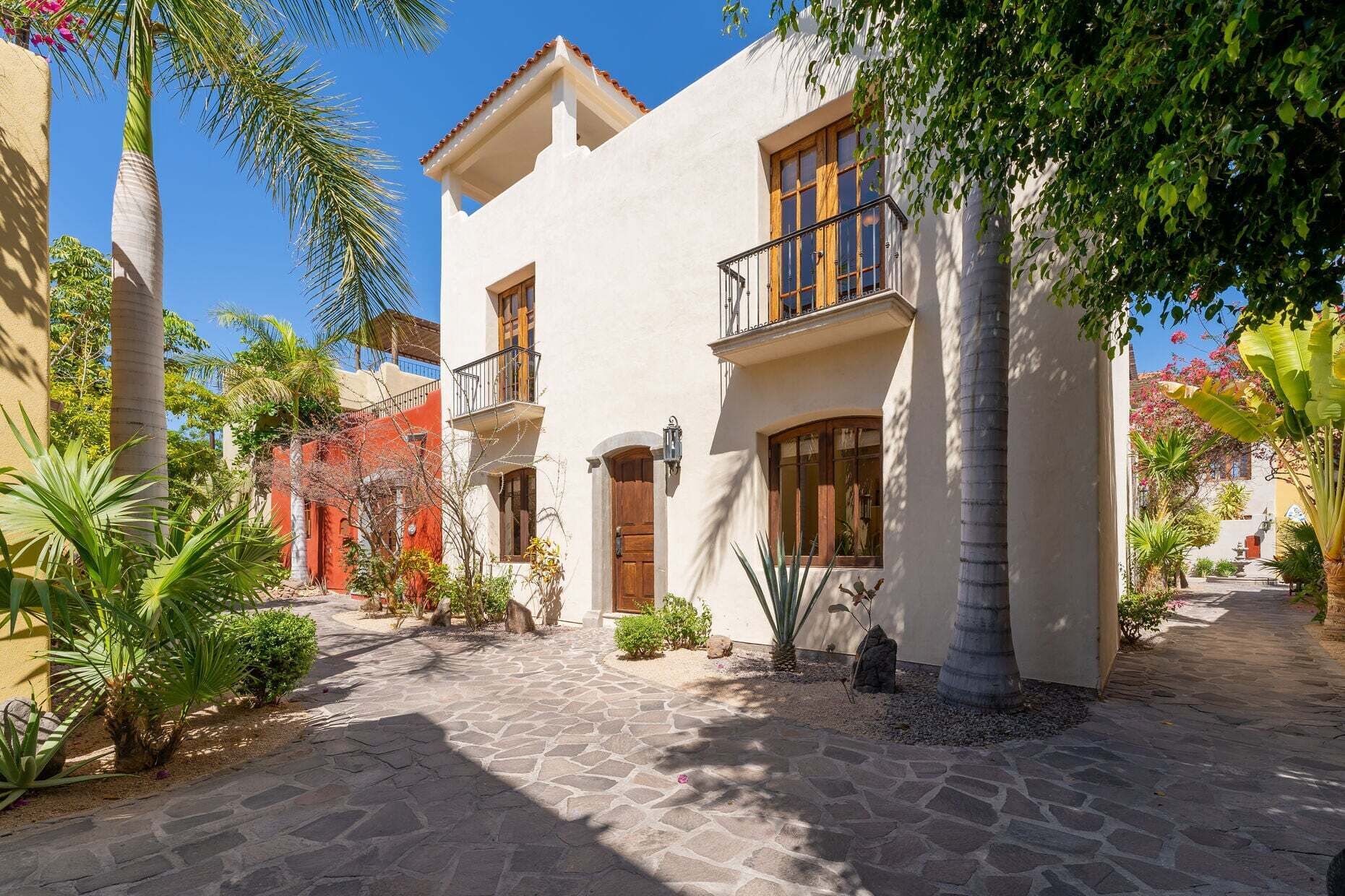 A two-story building with a stone-paved courtyard, surrounded by tropical plants and palm trees.