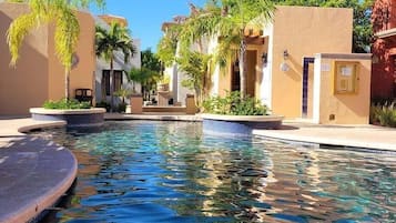 Un área de piscina con una piscina azul cristalina, rodeada de edificios con paredes color beige y palmeras.