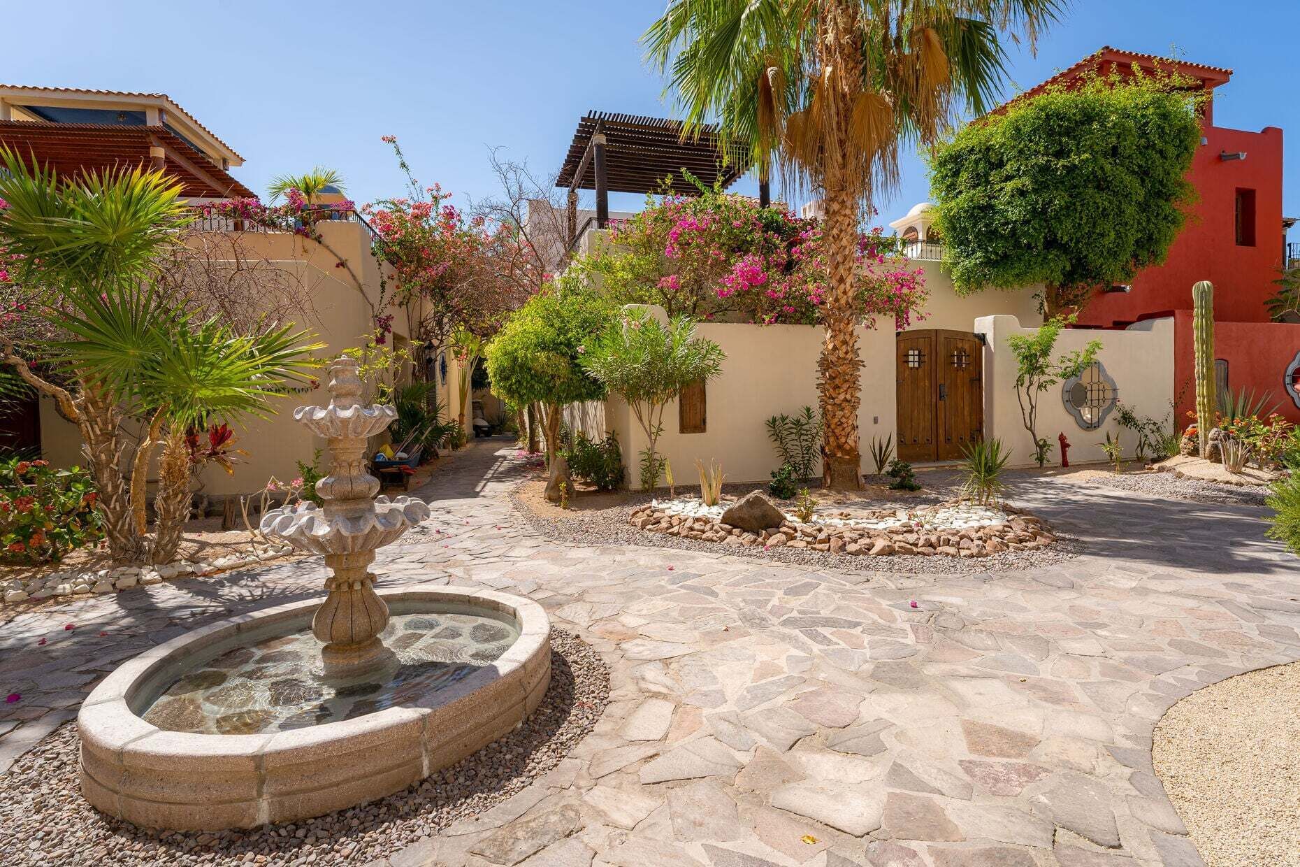 Un patio con una fuente de piedra, palmeras y plantas con flores.