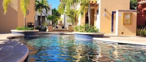 A pool area with a clear blue pool, surrounded by buildings with beige walls and palm trees.