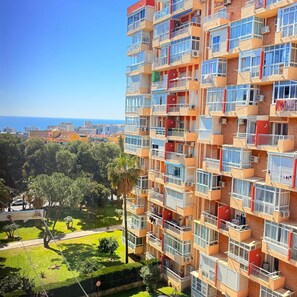 Ein mehrstöckiges Wohngebäude mit Balkonen, Blick auf das Meer und einer Grünfläche mit Bäumen.