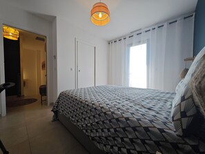 Ein Schlafzimmer mit einem gemusterten Bettbezug, einer Pendelleuchte, einer Tür und einem Fenster mit Vorhängen.