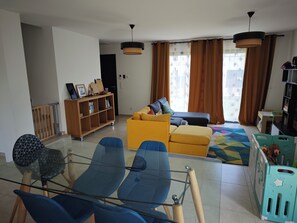 Ein Wohnzimmer mit einer Couch, Sesseln, einem Glastisch, einem Bücherregal und einem Spielbereich.