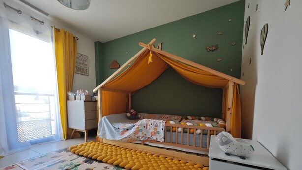 Huoneessa on sänky puisen katoksen alla, pieni valkoinen kaappi, keltainen neulottu matto ja kuviollinen lattiamatto.