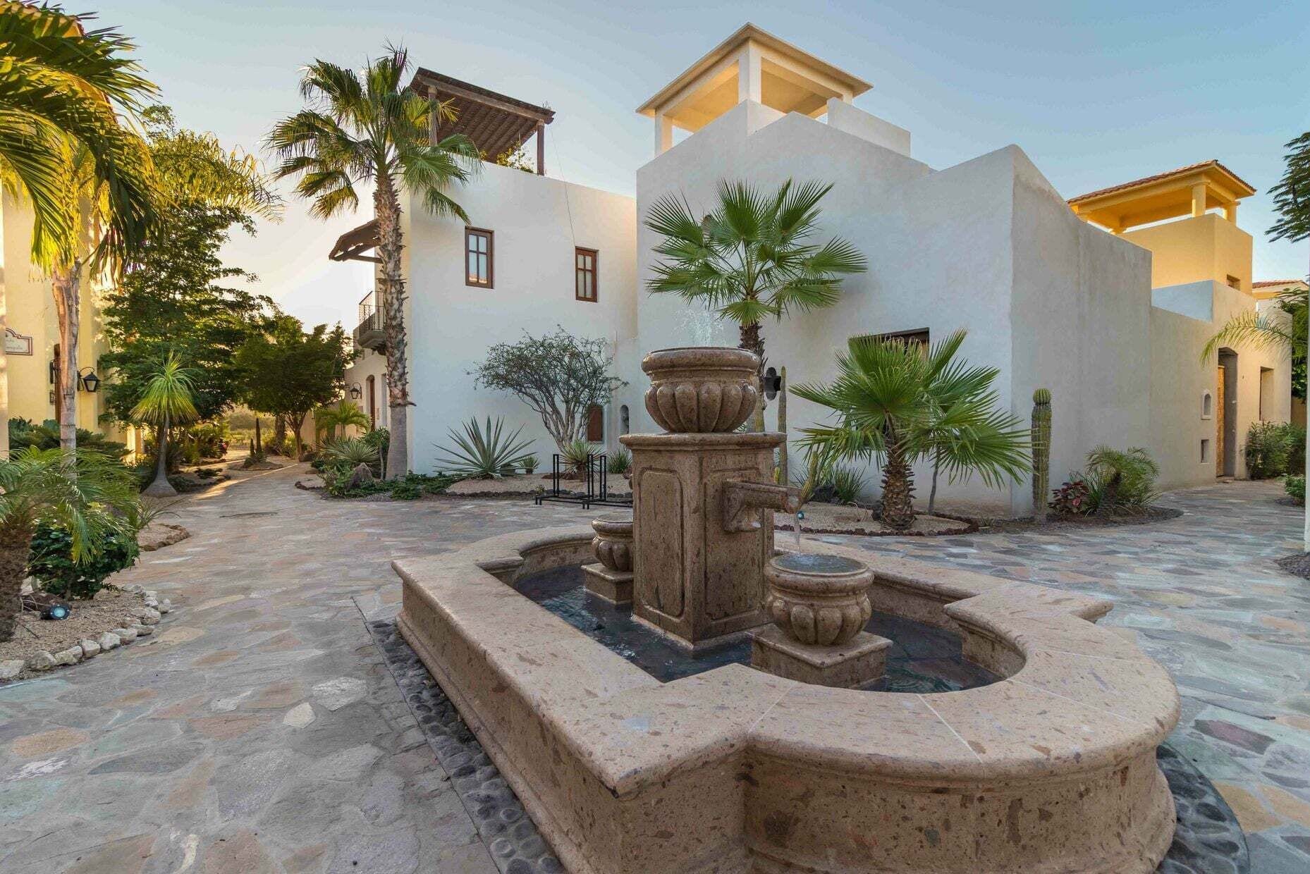 Un patio con una fuente, palmeras y un edificio de techo plano y paredes blancas.