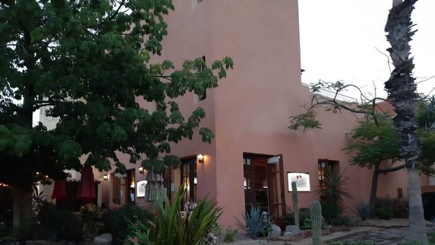 Un bâtiment à la façade rose, avec un espace extérieur aménagé avec des tables et des chaises, des parasols rouges et des cactus dans le jardin.