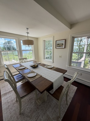 Comedor con una mesa de madera dispuesta para cuatro personas, sillas blancas, una araña y ventanas grandes con vistas a árboles y al cielo.