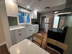 A modern kitchen with white cabinets, a stainless steel refrigerator, a gas stove, and a window with a view of trees. - Cozy 2-bedroom house in tranquil Chesterton with AC (Chesterton)