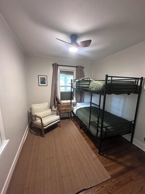 Una habitación con literas, una silla, una mesita pequeña, una ventana con cortinas y un ventilador de techo.