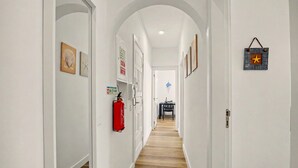 A hallway with white walls, wooden flooring, and framed artwork. There is a fire extinguisher mounted on the wall and a hanging decoration. - Apartment 40 Meters from the Beach with Balconies, Sea View, and Wi-Fi (Quarteira)