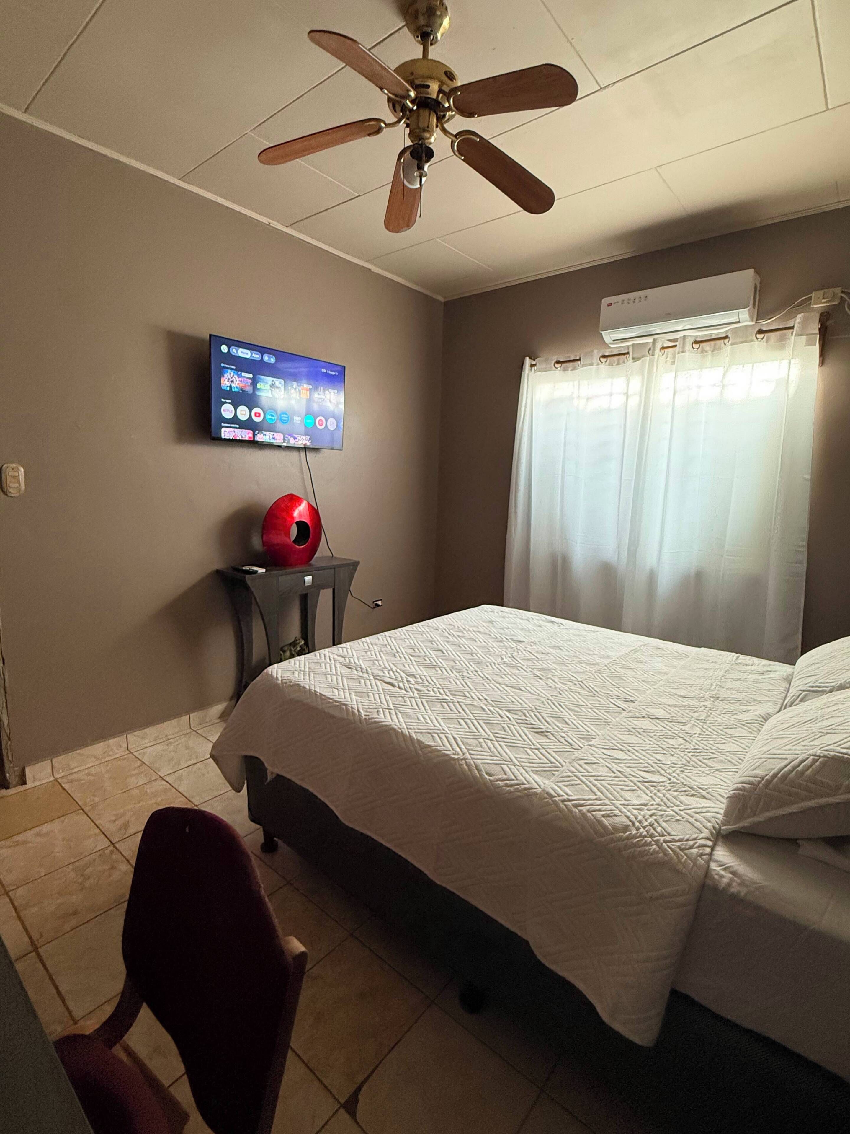 A hotel room with a bed, a ceiling fan, a TV mounted on the wall, and a window with curtains.