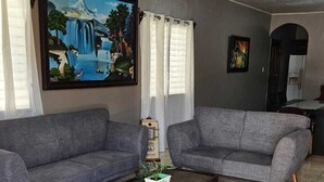 A living room with two grey sofas, a coffee table with a plant, a painting on the wall, and a kitchen area in the background. - Toronjal II Executive Rooms (La Ceiba)