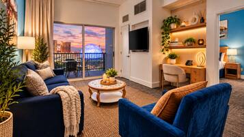 Une chambre d’hôtel moderne dotée d’un balcon, d’une télévision, d’un bureau avec une chaise et d’un coin salon avec des canapés et une table basse.