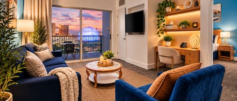 A modern hotel room with a balcony, a TV, a desk with a chair, and a seating area with sofas and a coffee table.