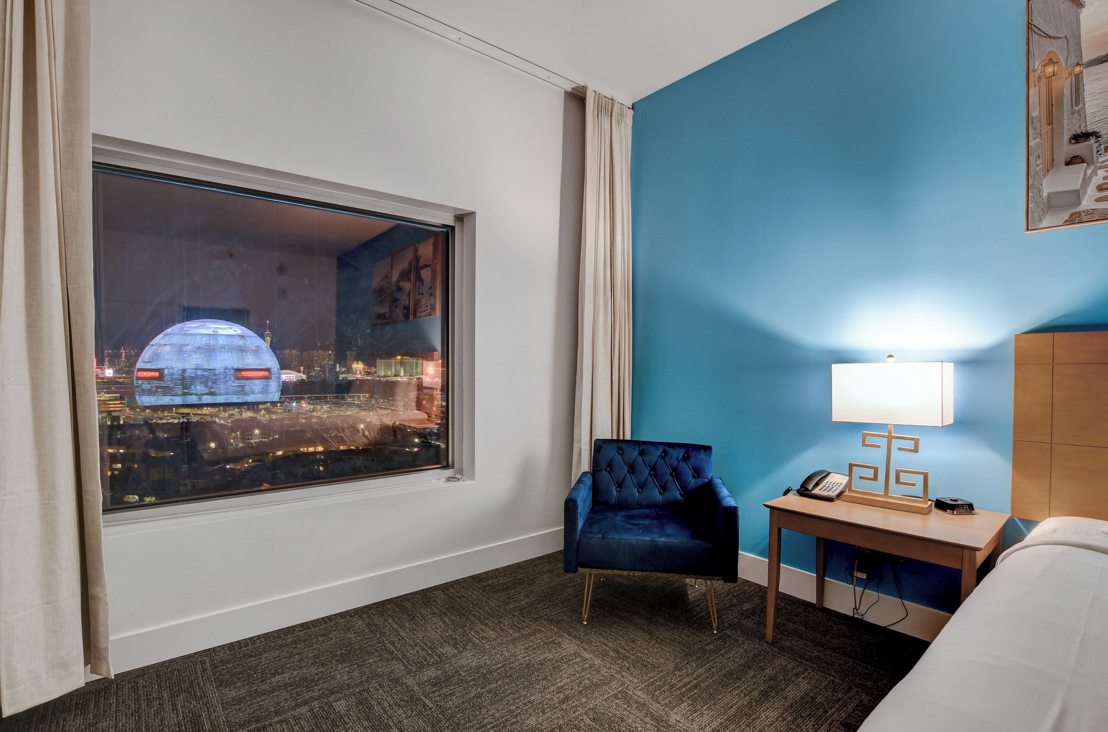 A hotel room with a large window, a blue chair, a bedside table with a lamp, and a bed.