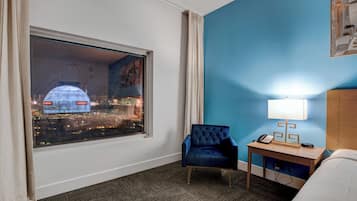 A hotel room with a large window, a blue chair, a bedside table with a lamp, and a bed.