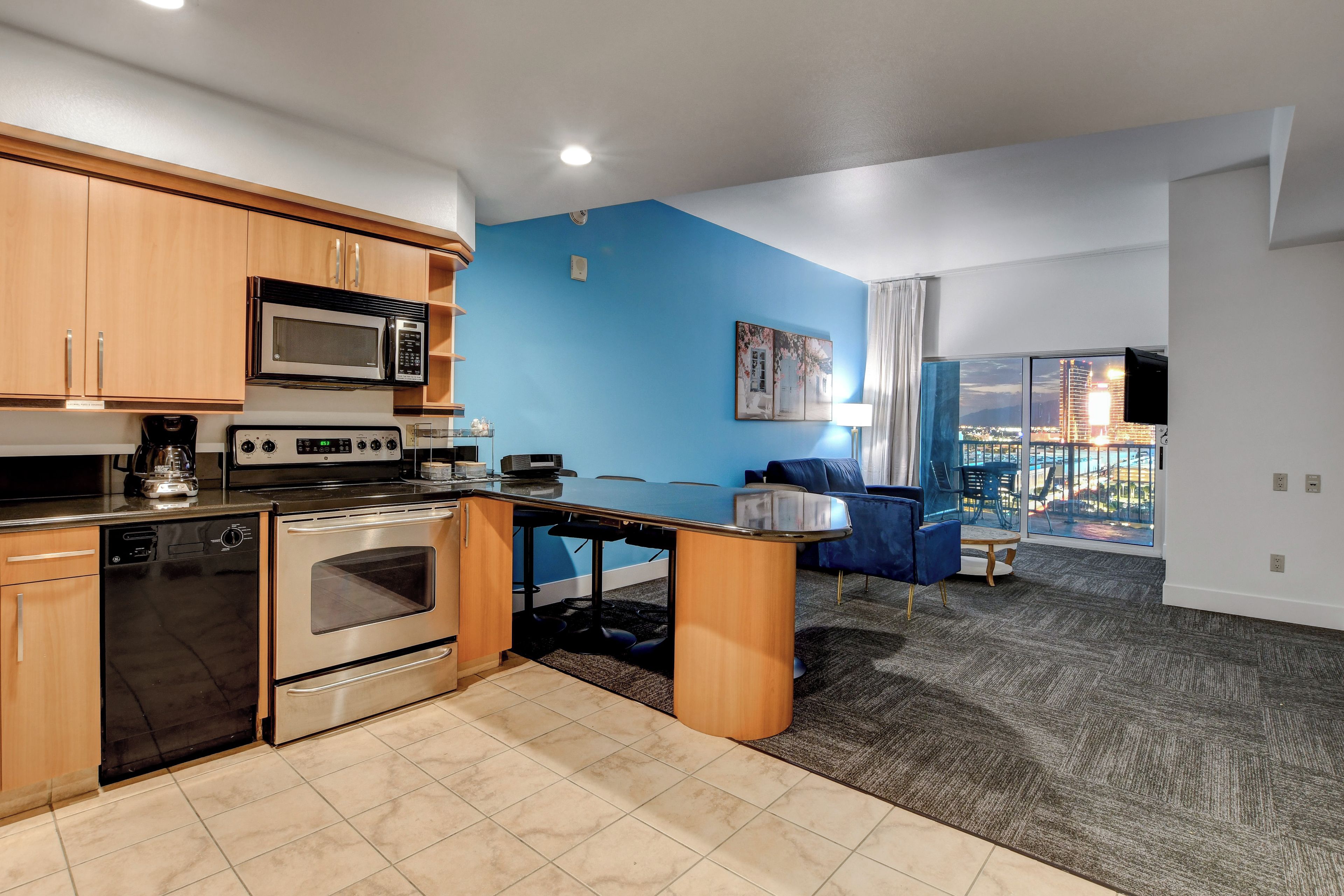 A modern kitchen with a microwave, stove, and dishwasher. There is a dining area with a table and chairs, and a living room with a blue wall and a large window.