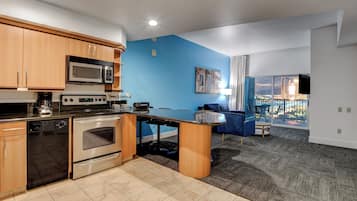 A modern kitchen with a microwave, stove, and dishwasher. There is a dining area with a table and chairs, and a living room with a blue wall and a large window.