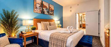 A hotel room with a large bed, a bedside table with a lamp, a chair, a potted plant, and a wall with three framed pictures.