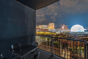 Un balcón con mesa y sillas, con vistas a un paisaje urbano con edificios iluminados y una estructura en forma de cúpula.