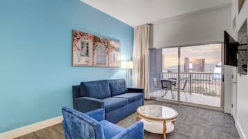 A hotel room with a blue wall, a sofa, a chair, a small table, and a balcony with a view of buildings.