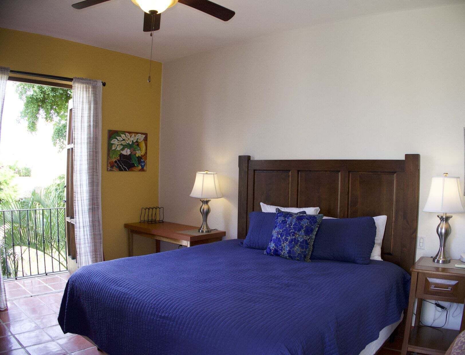 Un dormitorio con cama, dos lámparas de noche, una mesita de madera, un cuadro en la pared y un balcón con vistas a zonas verdes.