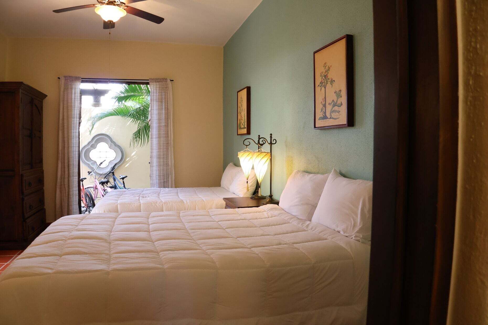 Un dormitorio con cama, ventilador de techo, mesita de noche con lámpara y una bicicleta al fondo.