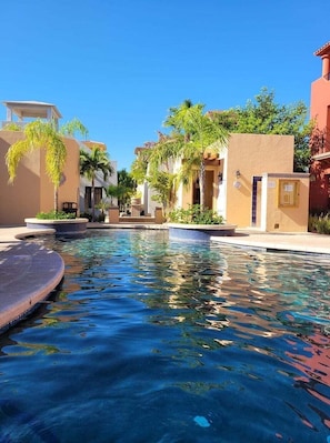 Una piscina con superficie de agua azul cristalina, rodeada de edificios de paredes color beige y palmeras.
