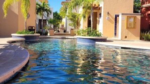 A pool area with a clear blue pool, surrounded by buildings with beige walls and palm trees. - Casa del Gallo-home in Loreto Bay- Baja Reimagined! Steps From Community Pool (Loreto Bay)