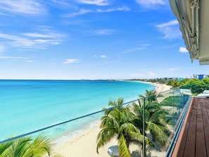Blick auf einen Strand von einem Balkon aus, mit blauem Himmel, Palmen und einem Holzboden.