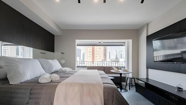 Ett modernt hotellrum med en stor säng, en tv, ett litet bord och utsikt över staden.