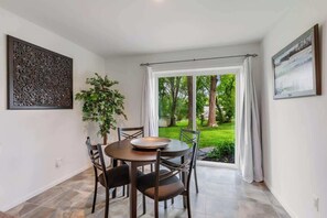 Ein Essbereich mit Tisch und Stühlen, einer Topfpflanze und Blick auf einen Garten durch eine Glastür.