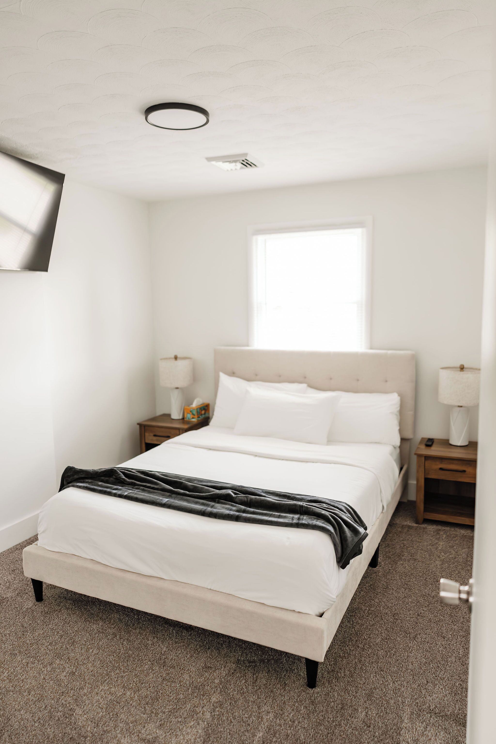 A bedroom with a bed, nightstands, a window, and a wall-mounted TV.