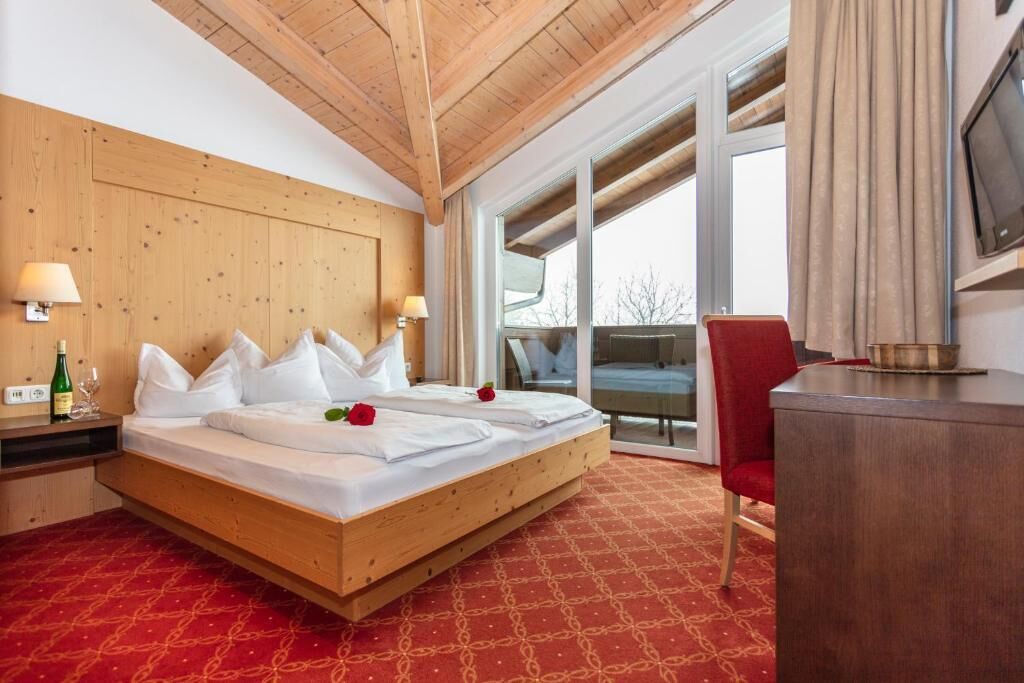 Habitación de hotel con techo de madera, una cama grande con ropa de cama blanca, dos lámparas de noche, una silla roja y un televisor en la pared.