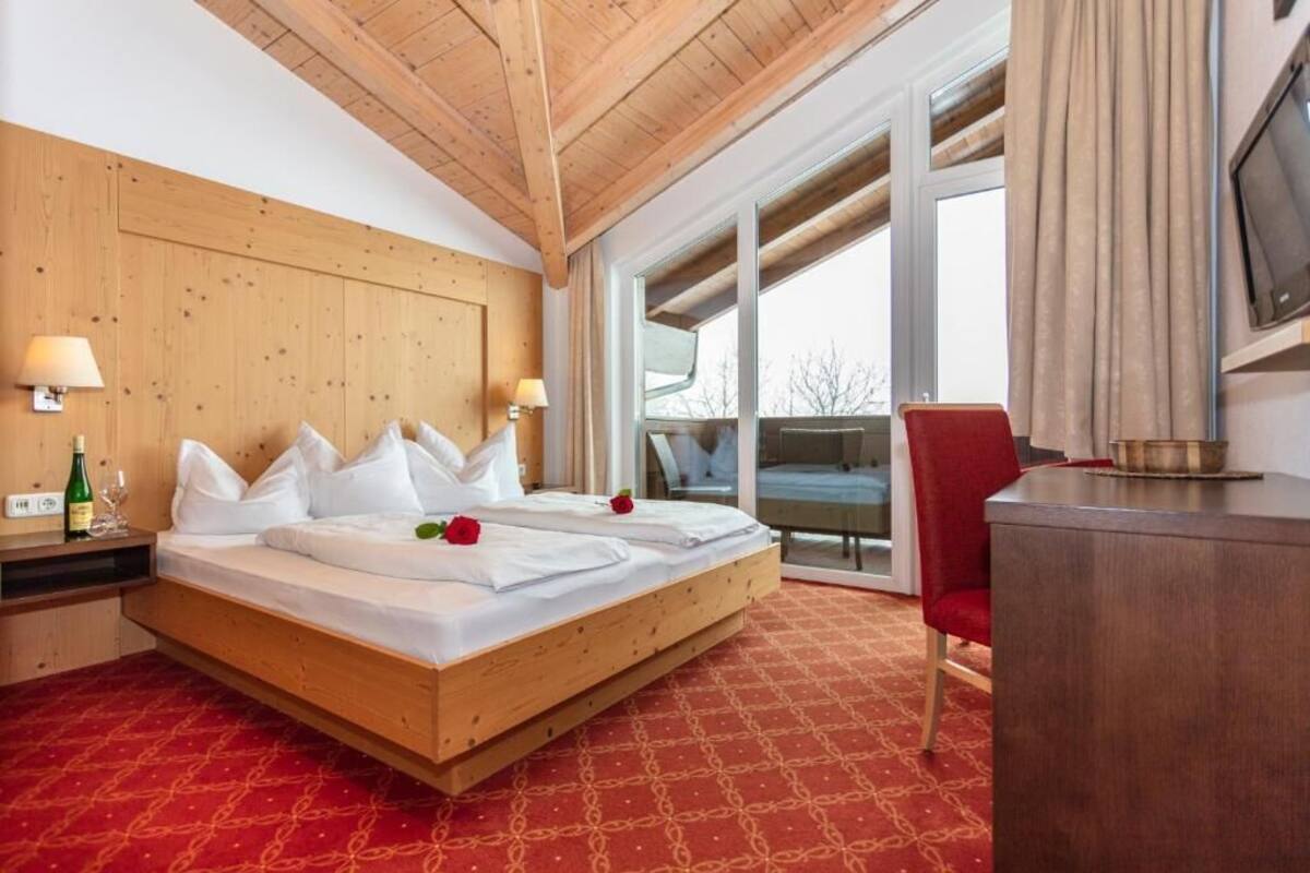 Habitación de hotel con techo de madera, una cama grande con ropa de cama blanca, dos lámparas de noche, una silla roja y un televisor en la pared.