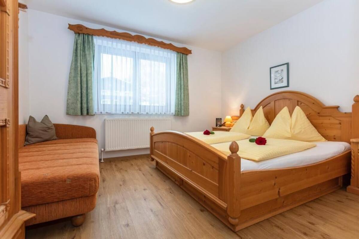 Un dormitorio con cama y sofá de madera, ventana con cortinas, radiador y un cuadro enmarcado en la pared.