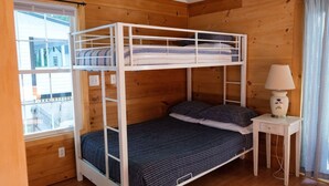 A bunk bed with a side table and lamp, wooden walls, a window, and a view of buildings outside.