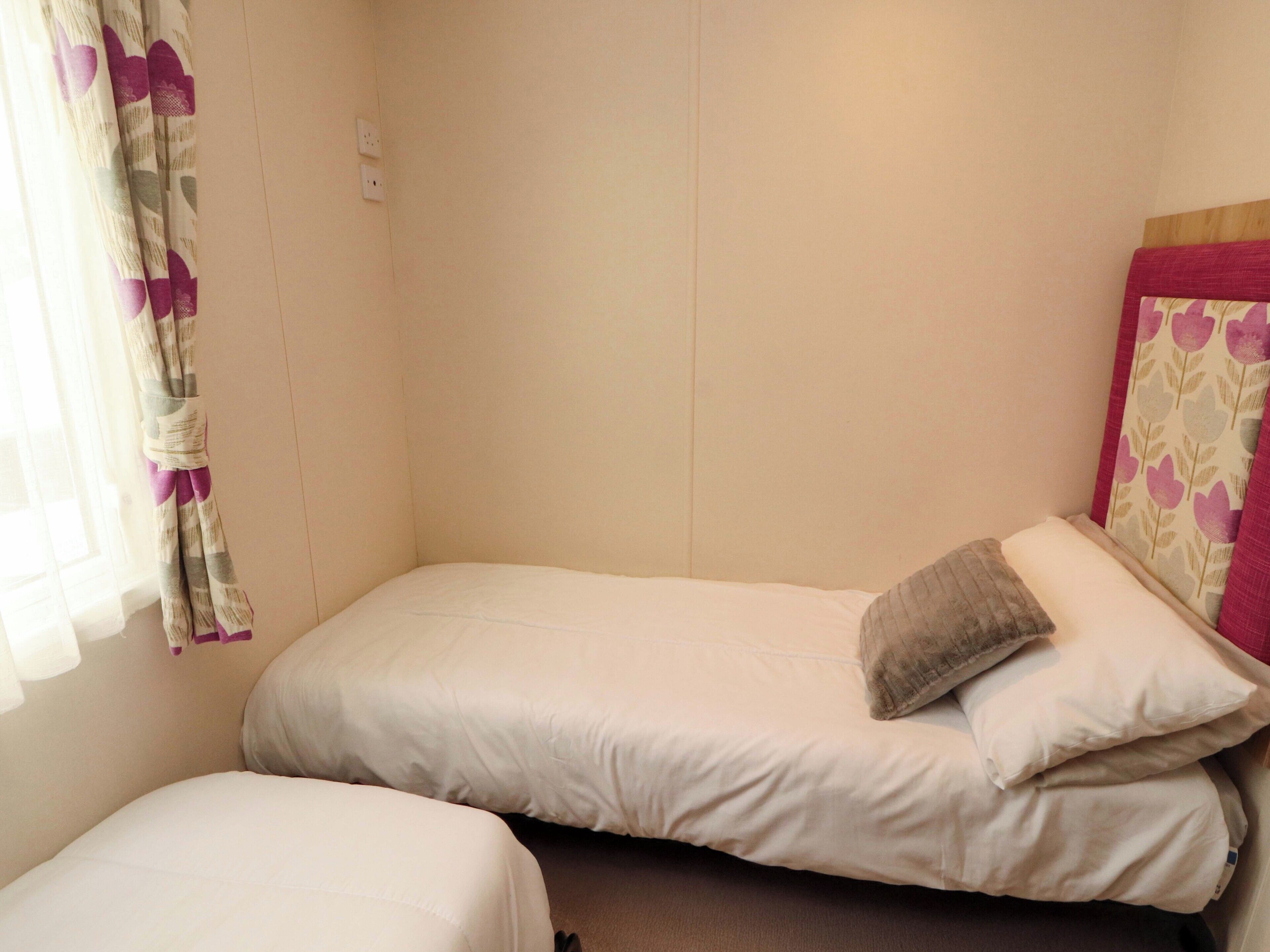 A small, single-bed room with a plain wall, a window with curtains, and a bedside table.
