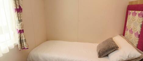 A small, single-bed room with a plain wall, a window with curtains, and a bedside table.