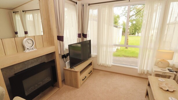 A living room with a fireplace, a television, and a view of a green lawn. - Daletree View (Skipton)