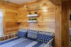 Un intérieur en bois avec une pancarte « BIENVENUE », un lit avec une literie à motifs et une étagère avec des plantes.