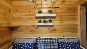 Ein Holzinnenraum mit einem 'WELCOME'-Schild, zwei Pflanzenregalen und drei gemusterten Kissen auf einer Bank.