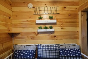 Un intérieur en bois agrémenté d’une pancarte « BIENVENUE », de deux étagères ornées de plantes et de trois coussins à motifs disposés sur un banc.