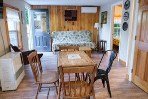 Ein gemütliches Zimmer mit Holzboden, einer Couch, einem Esstisch mit Stühlen, einem kleinen Heizgerät und einer Tür zu einem anderen Raum.
