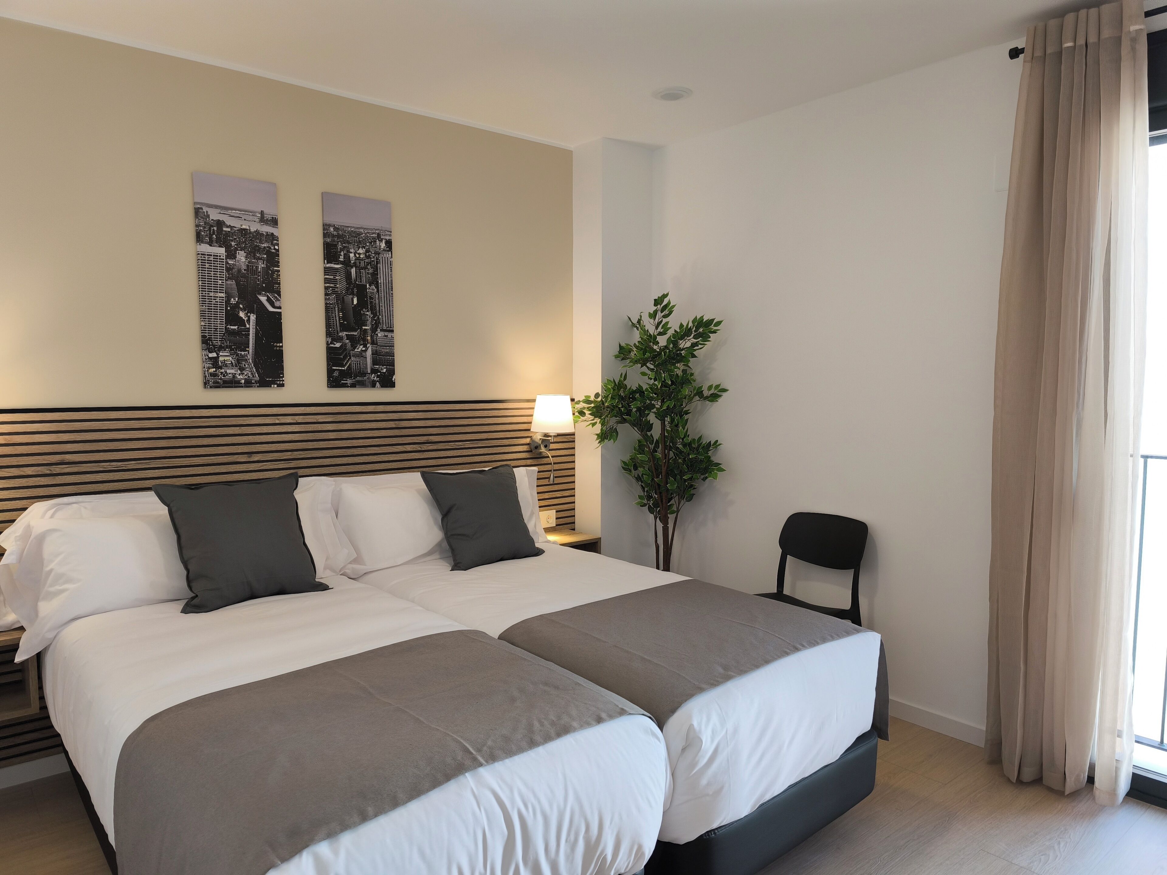 A double bed with two pillows, a nightstand with a lamp, a potted plant, and a chair.