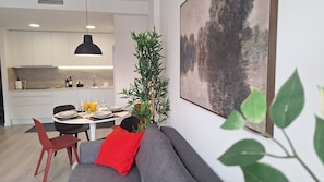 Una cocina moderna con armarios blancos, un comedor con una mesa redonda y un sofá con un cojín rojo.