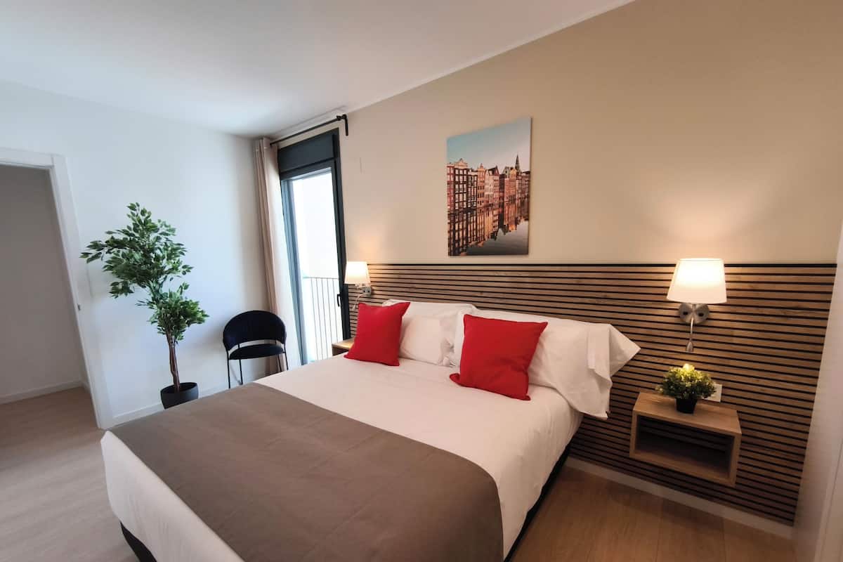 Hotellihuone, jossa on suuri sänky, ruukussa oleva kasvi, tuoli, yöpöytä lampun kanssa ja seinällä kaupunkimaisemaa esittävä taulu.