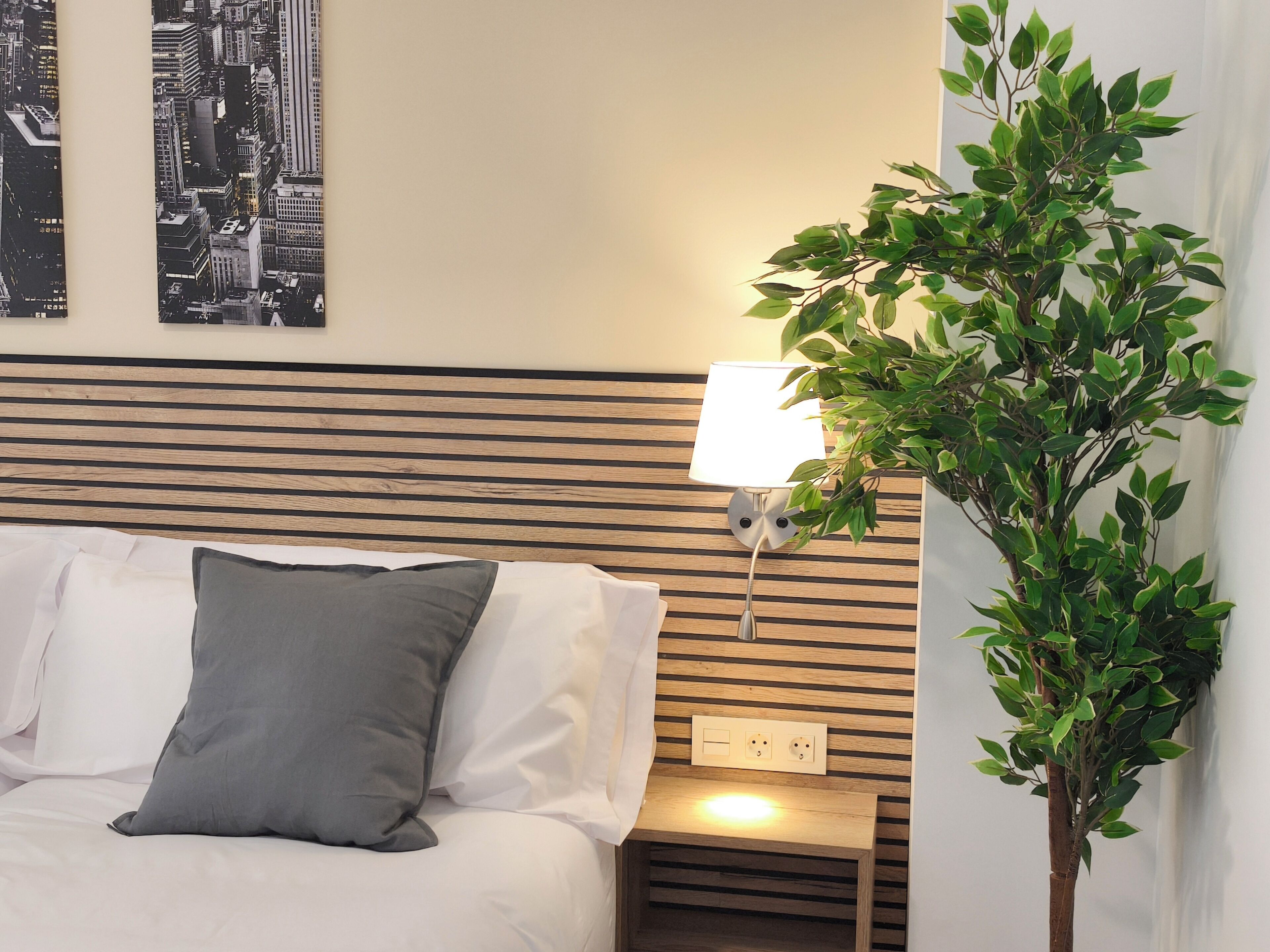 Un lit bien fait, avec des oreillers blancs et gris, une tête de lit en bois, une lampe fixée au mur et une plante en pot.