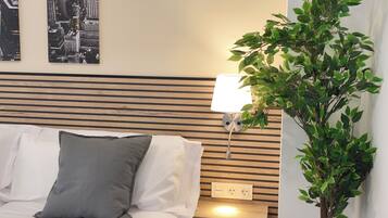 Un lit bien fait, avec des oreillers blancs et gris, une tête de lit en bois, une lampe fixée au mur et une plante en pot.